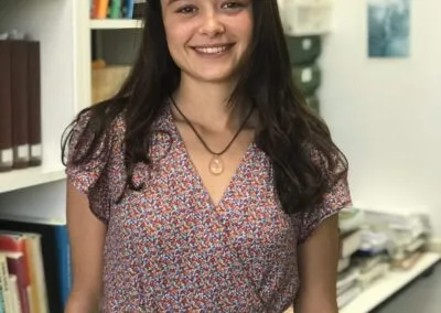 Una mujer joven sonríe a la cámara con blusa estampada y colgante, en una oficina con estanterías y carpetas al fondo.