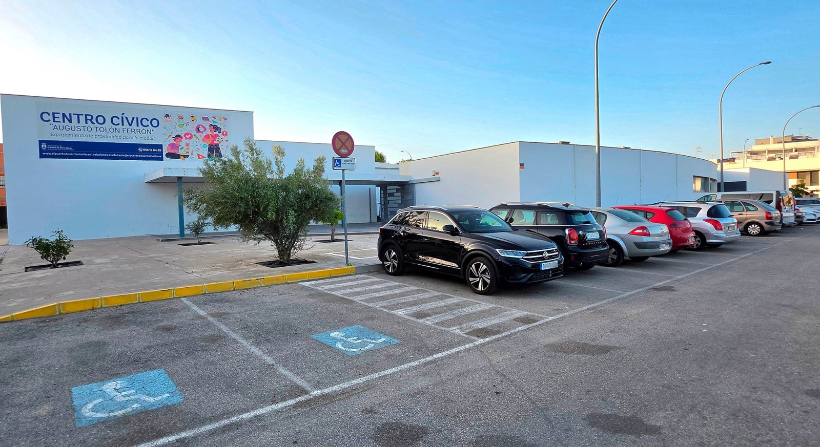 Fachada blanca de un centro cívico con cartel azul, aparcamiento con coches y plazas para discapacitados.