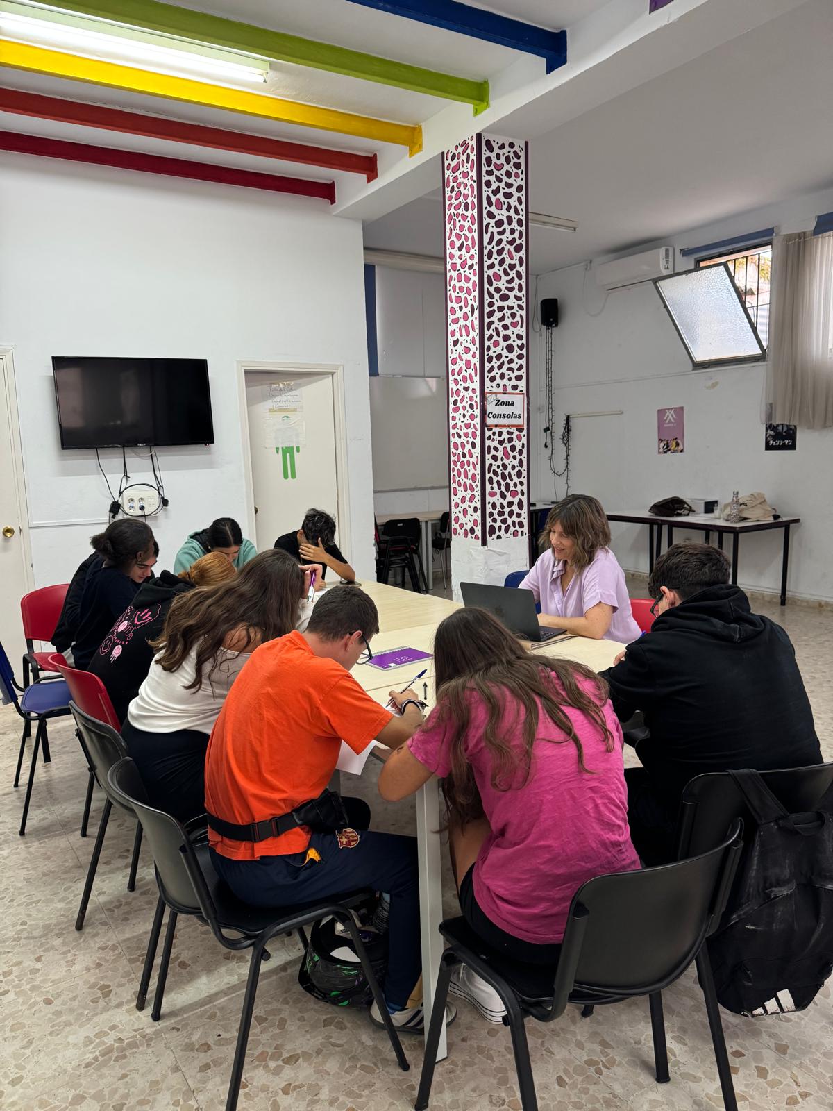 Varias personas jóvenes sentadas alrededor de una mesa larga escriben y trabajan con un portátil en una sala con vigas de colores.