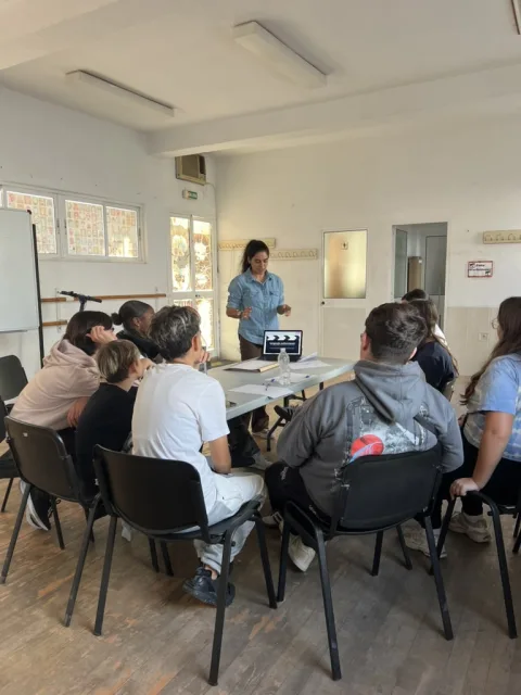 Una mujer de pie habla ante un grupo de jóvenes sentados en sillas alrededor de una mesa con un portátil, en un aula luminosa.