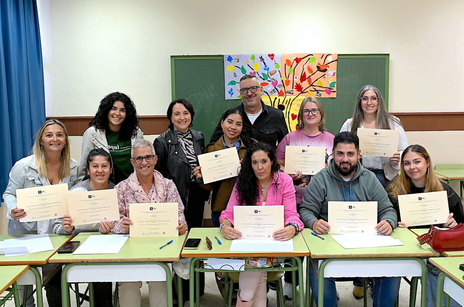 Un grupo de personas posa sonriente en un aula, de pie y sentadas tras pupitres, sosteniendo diplomas.