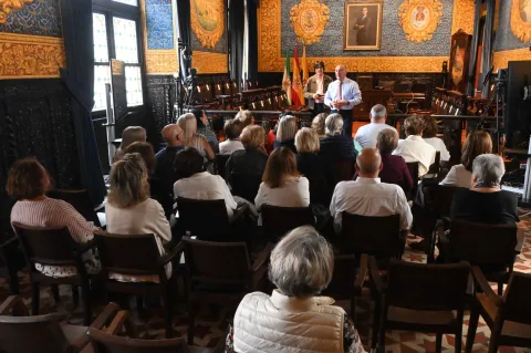 Un grupo de personas sentadas de espaldas escucha a dos hombres de pie en una sala con paredes decoradas y banderas.