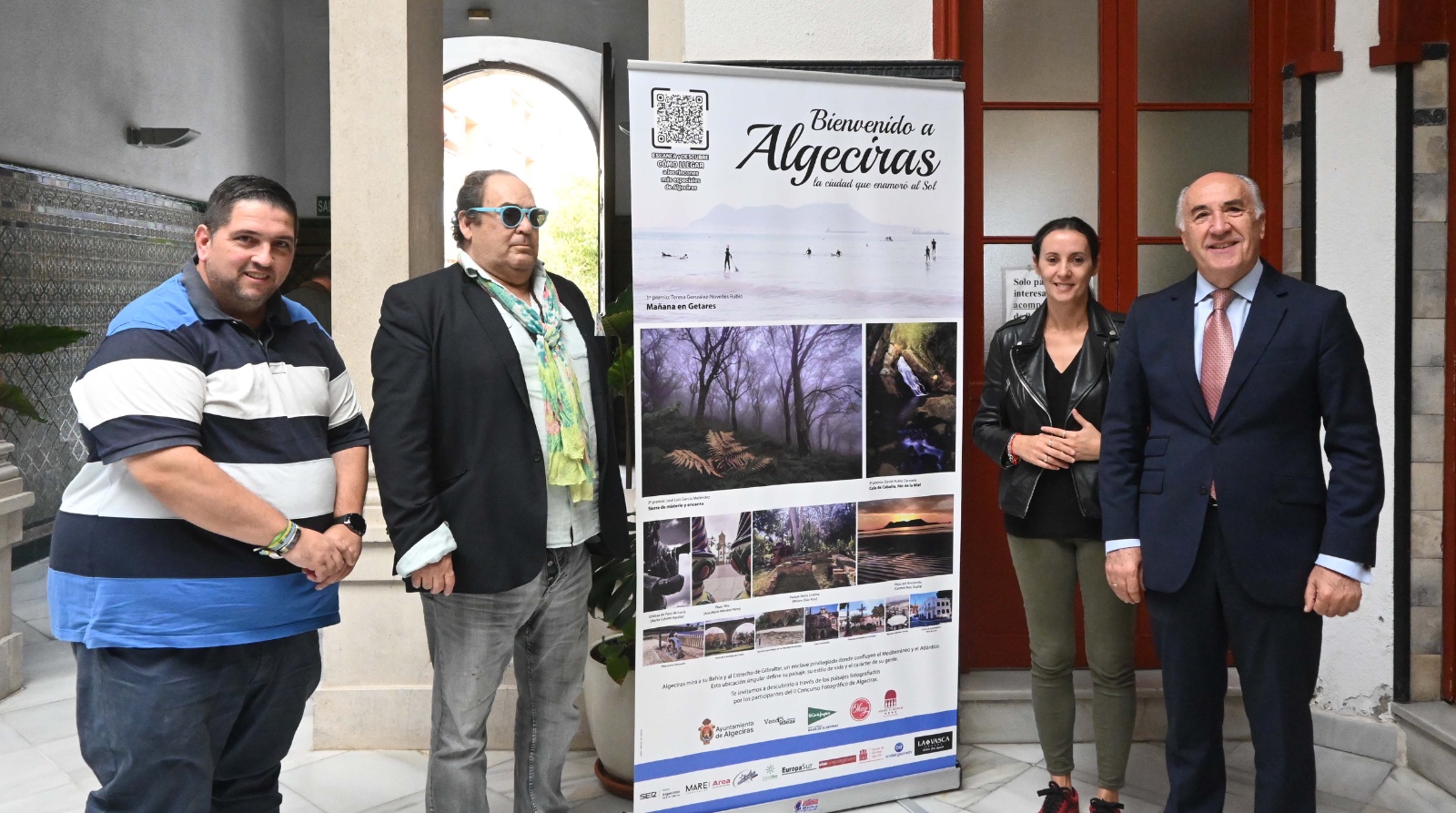 Cuatro personas posan junto a un cartel turístico enrollable sobre Algeciras en un patio interior.