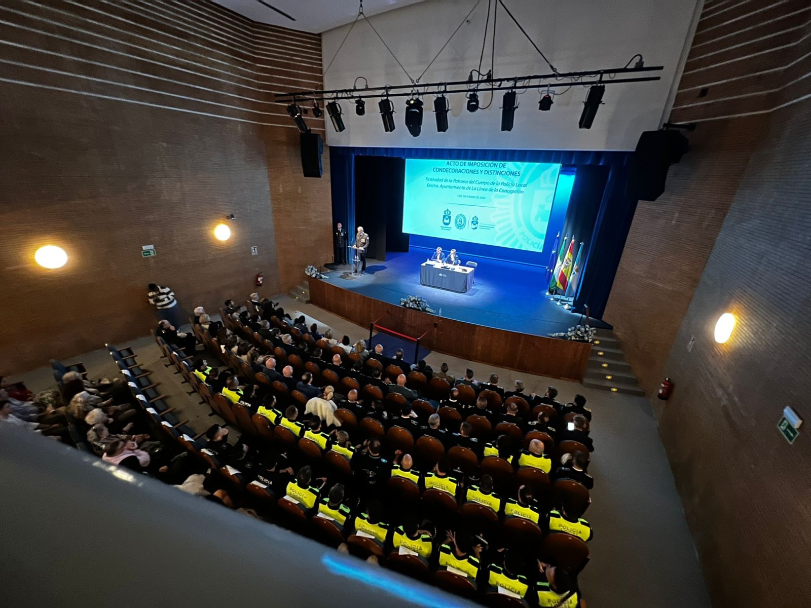 Vista elevada de un auditorio con público sentado, muchos con chalecos amarillos de policía, y una pantalla azul en el escenario.