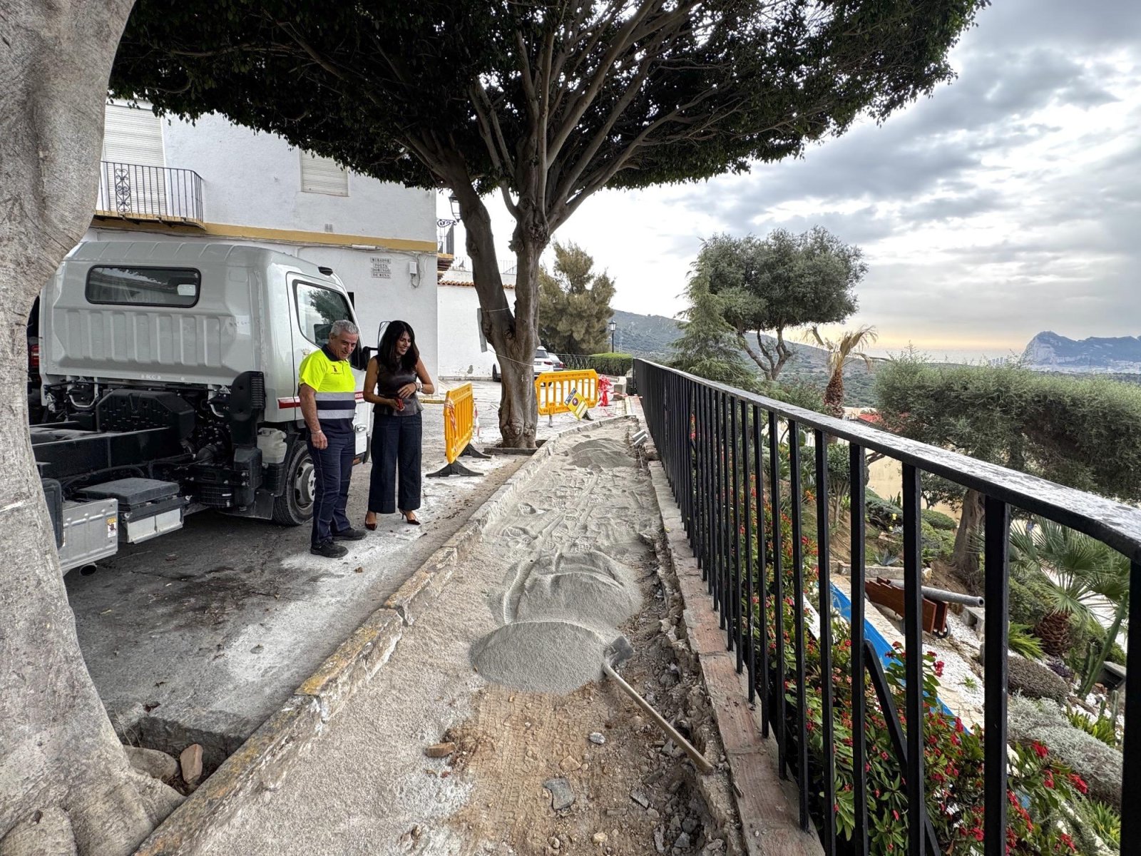 Un hombre con chaleco reflectante y una mujer junto a un camión, ante una acera en obras con arena y una barandilla.