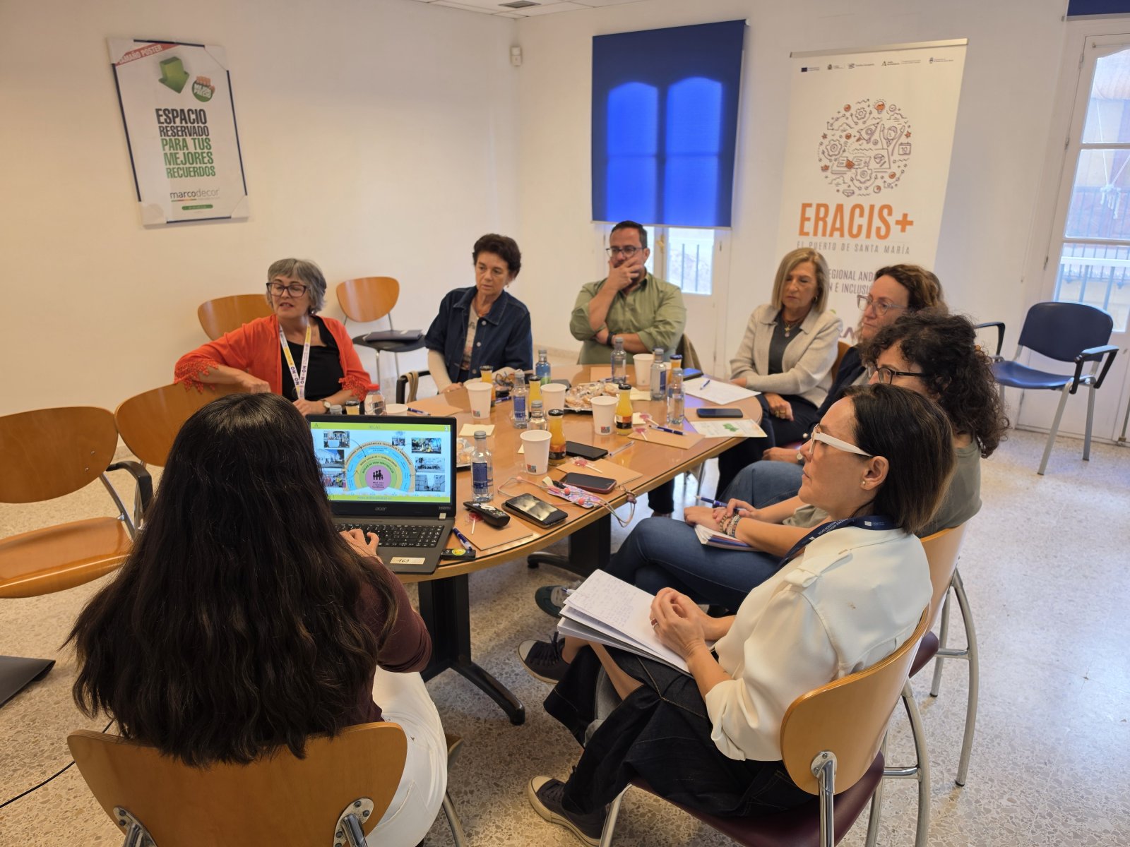 Siete personas sentadas alrededor de una mesa ovalada con un portátil, botellas y papeles, en una sala de reuniones con carteles.