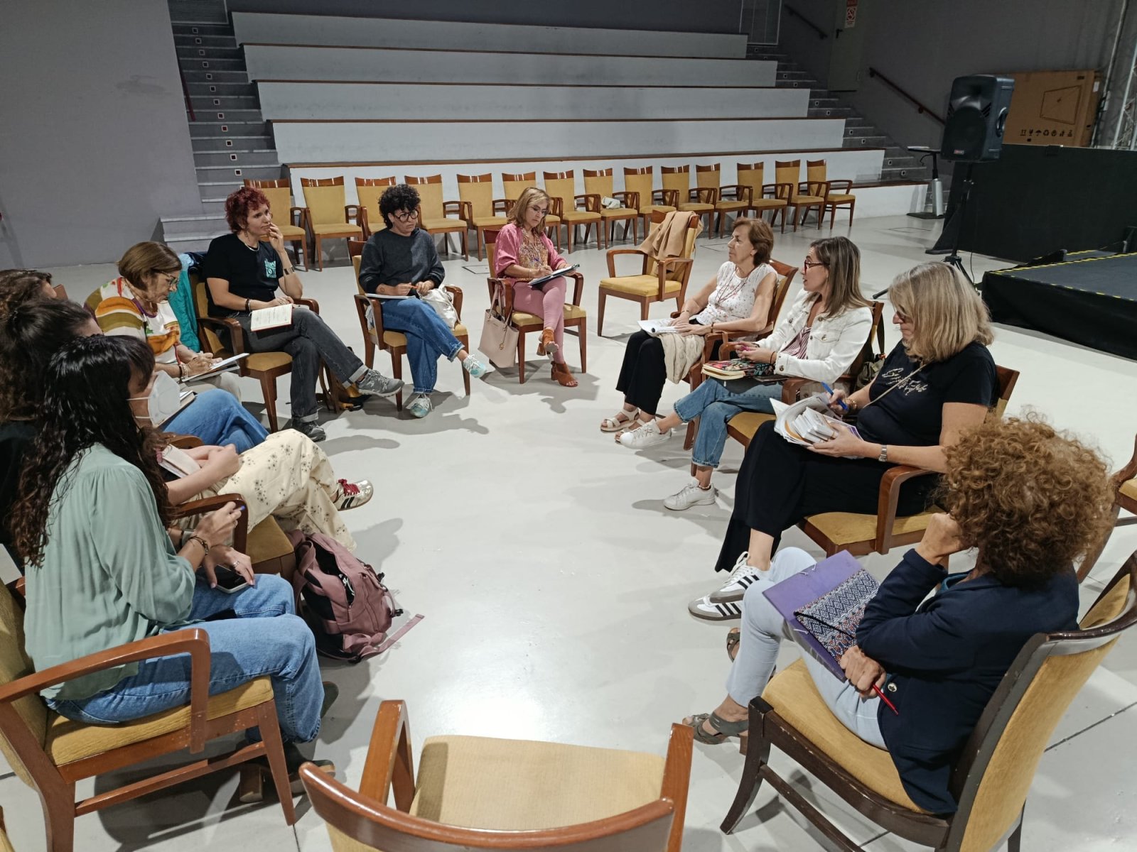 Un grupo de personas sentadas en círculo con sillas y cuadernos en una sala amplia con gradas al fondo.