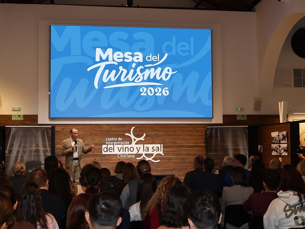 Un hombre habla con micrófono ante un público sentado, bajo una pantalla que dice Mesa del Turismo 2026.