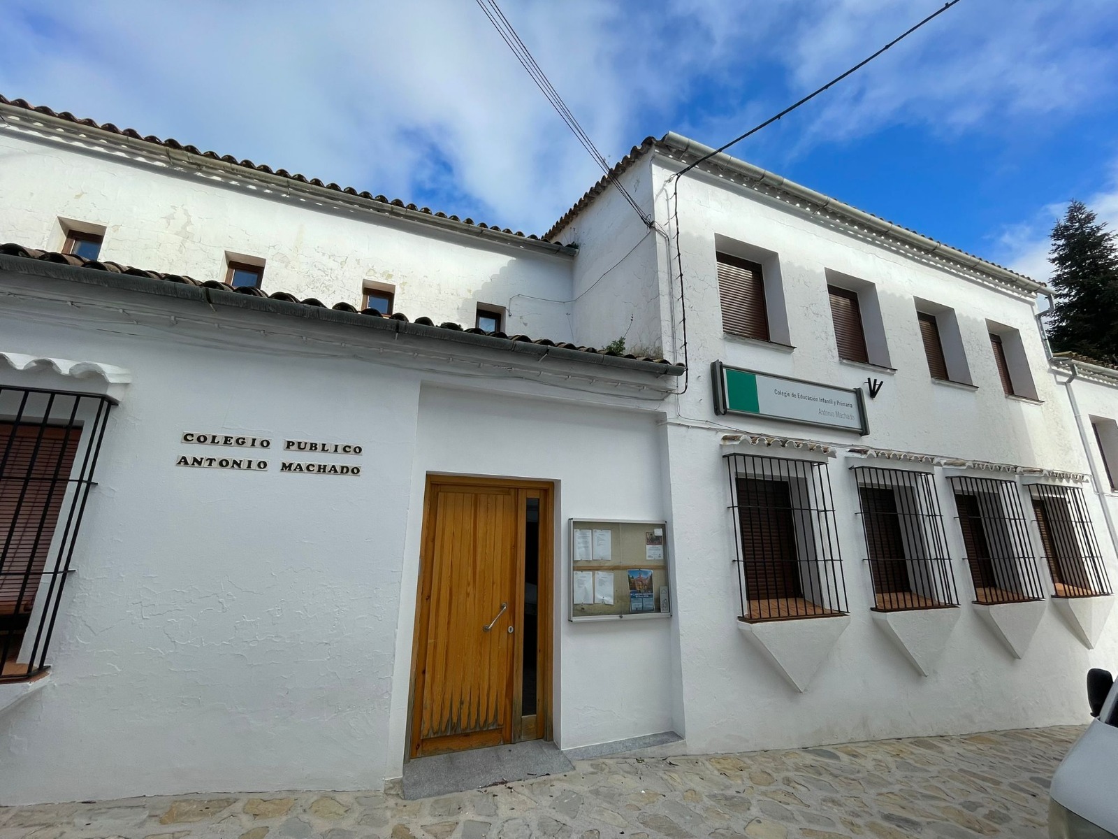 Fachada blanca de un colegio público con puerta de madera, ventanas enrejadas y un rótulo que dice Colegio Público Antonio Machado.