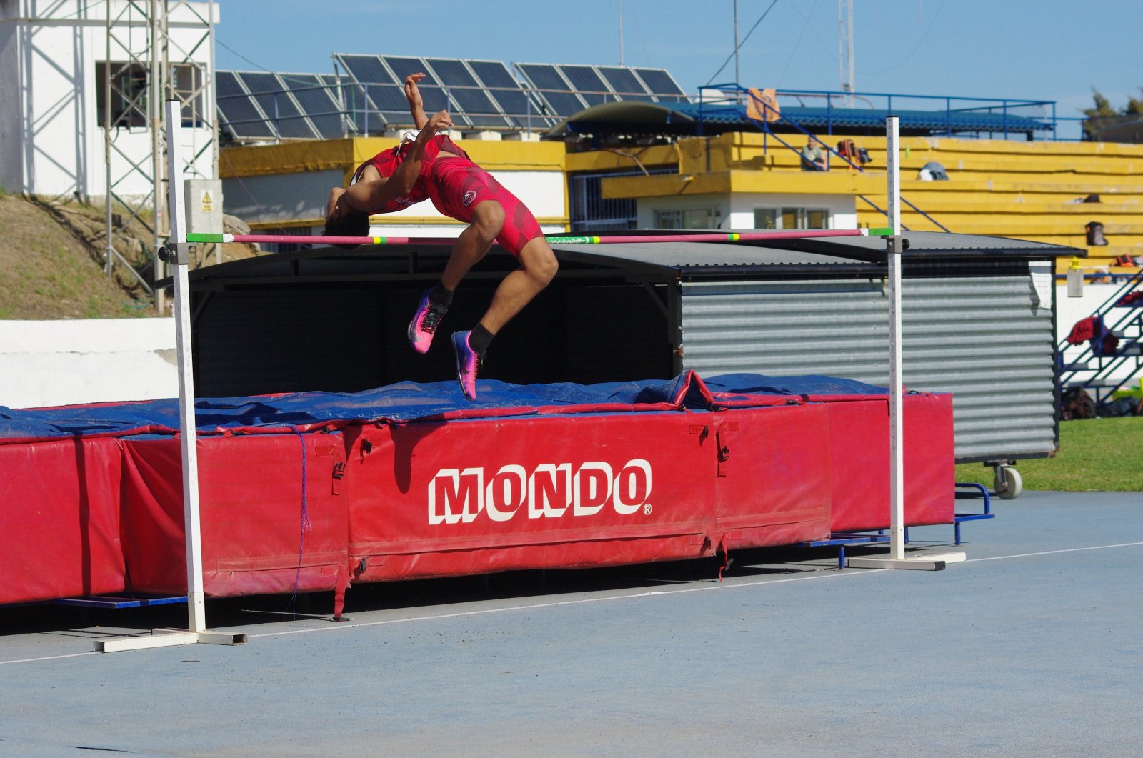 Un atleta con ropa rosa salta sobre el listón en una colchoneta roja con el rótulo Mondo, en una pista de atletismo.