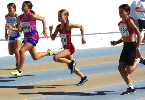 Cuatro corredoras jóvenes con dorsal compiten en una pista de atletismo azul y beige junto a un muro blanco.