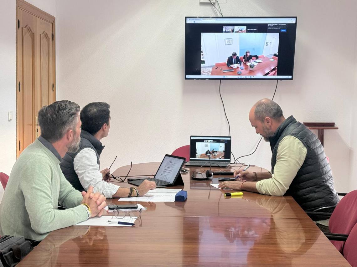 Tres hombres sentados ante una mesa de reuniones con portátiles, uno de ellos frente a una pantalla que muestra una videollamada.