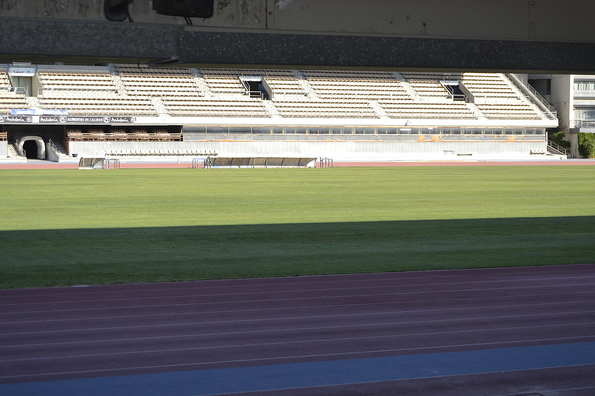 Campo de fútbol vacío con césped verde, pista de atletismo y gradas cubiertas sin público.