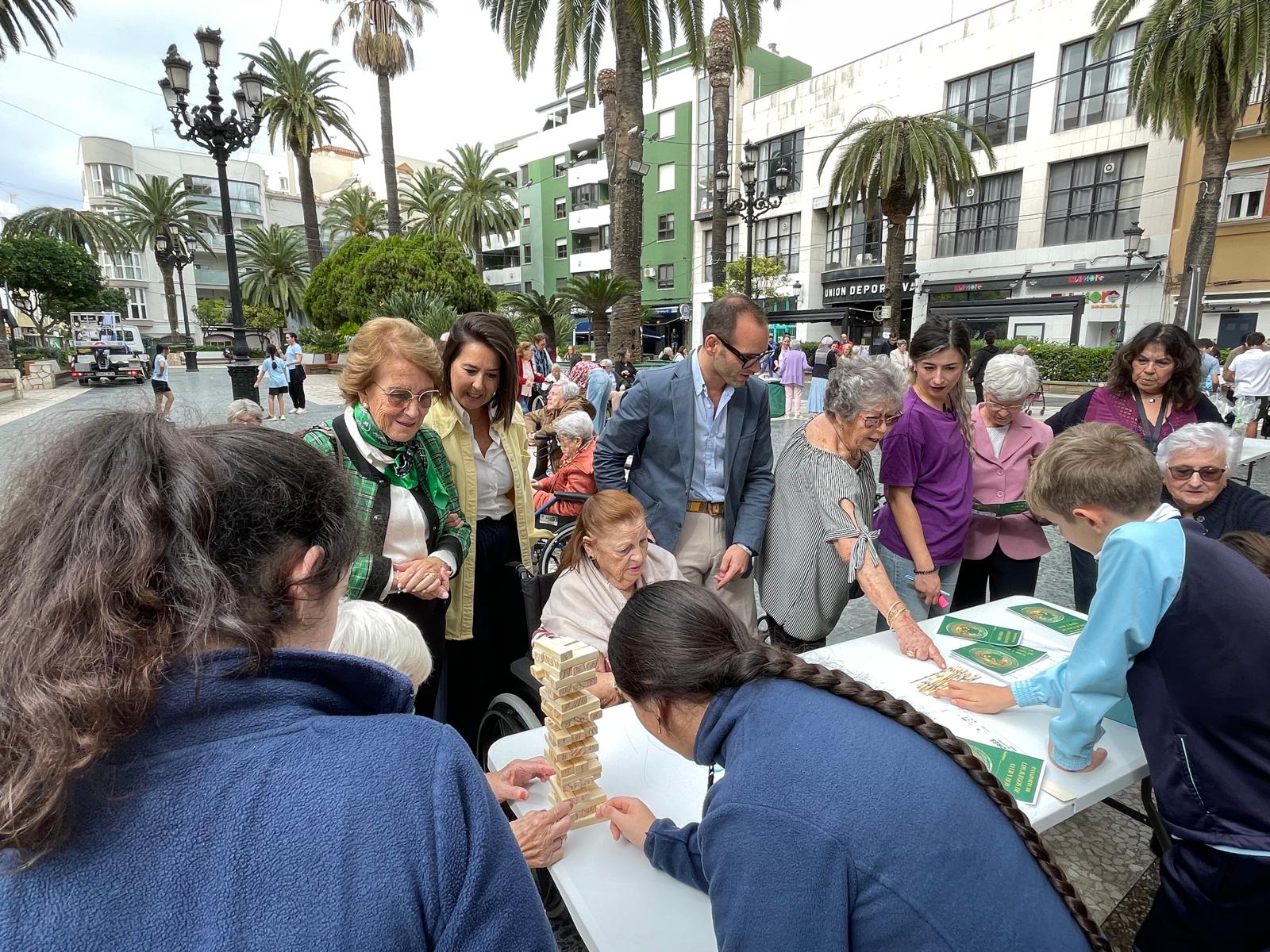 Personas mayores y jóvenes juegan alrededor de una mesa con una torre de bloques de madera en una plaza con palmeras.