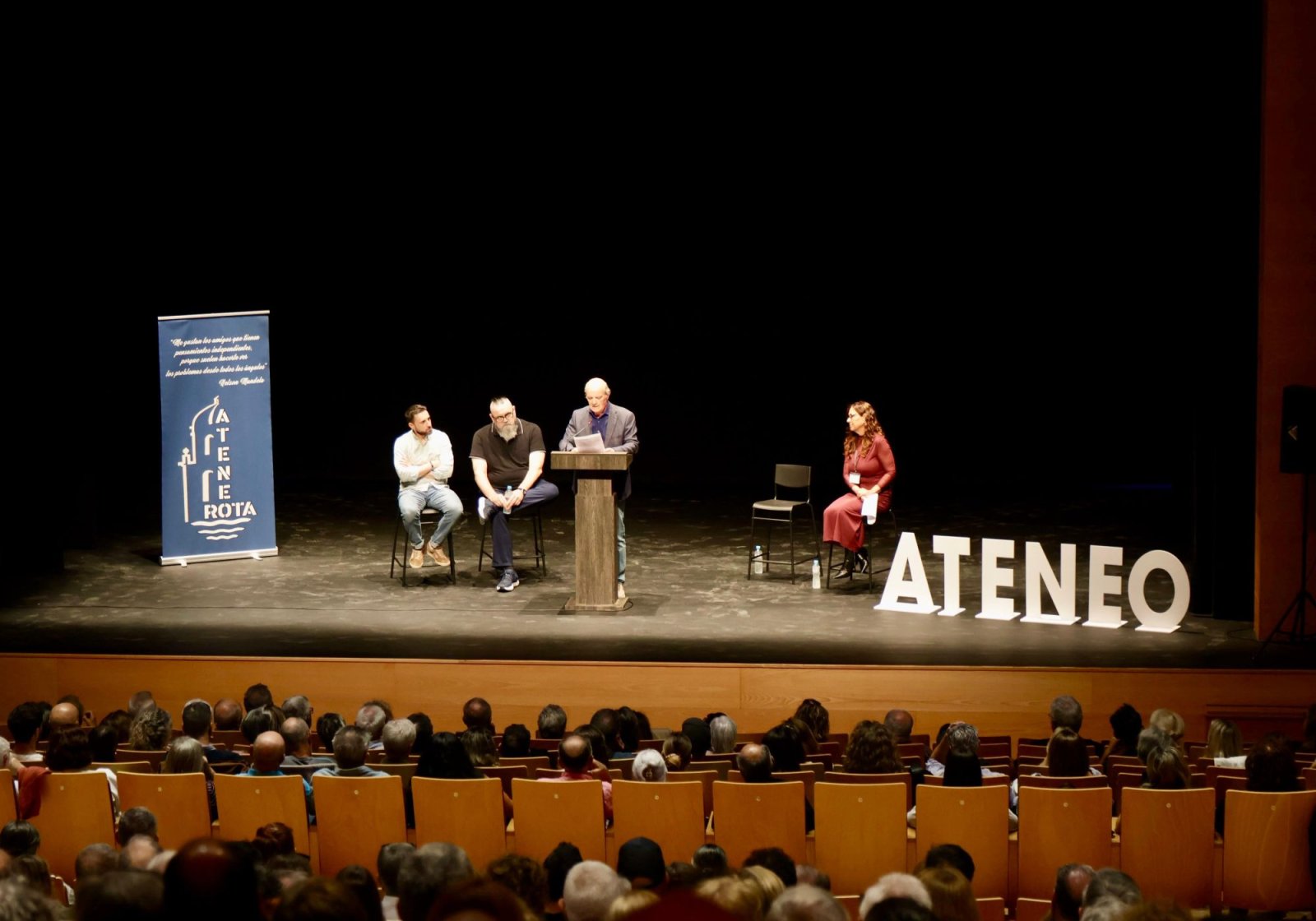 Cuatro personas en un escenario ante un público sentado, con un cartel que dice ATENEO ROTA.