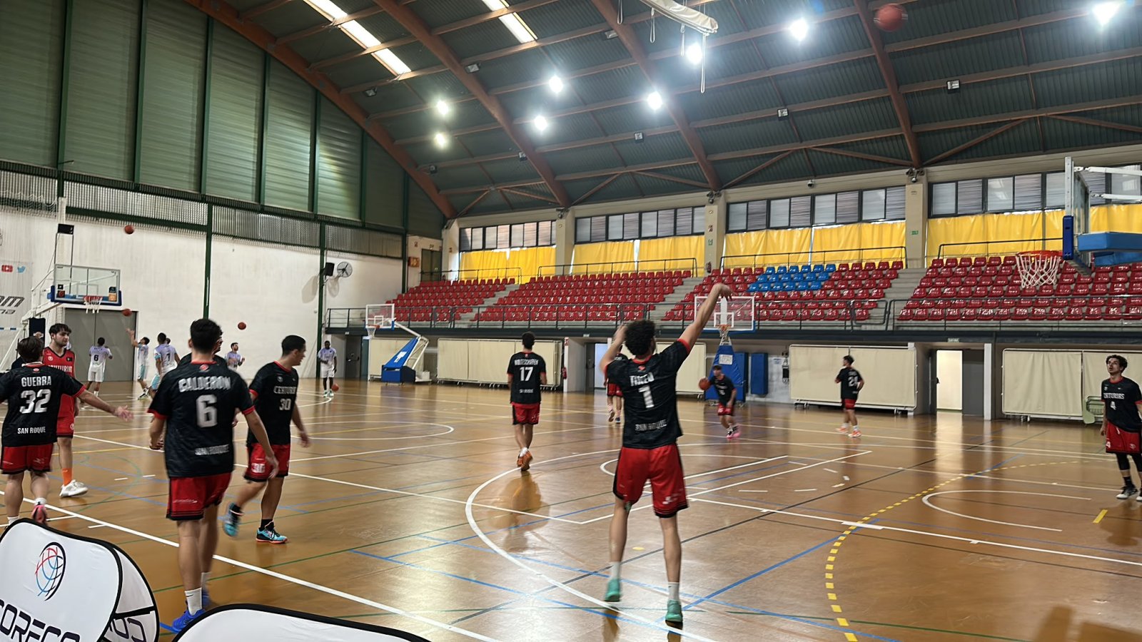 Jugadores de baloncesto con camisetas negras y rojas dispersos por la pista de un pabellón con gradas rojas al fondo.
