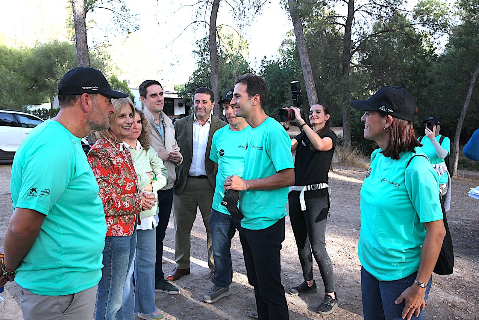 Un grupo de personas conversa de pie en un camino de tierra entre árboles, varias con camisetas verdes y gorras, y una graba con cámara.