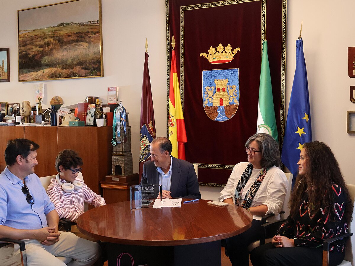 Cuatro personas conversan sentadas alrededor de una mesa ovalada en una sala con banderas y un escudo bordado en la pared.