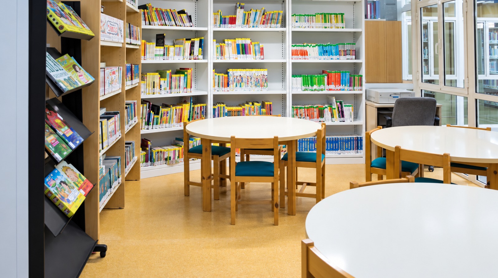 Estanterías blancas llenas de libros de colores, un expositor inclinado y mesas redondas con sillas de asiento azul en una sala luminosa.