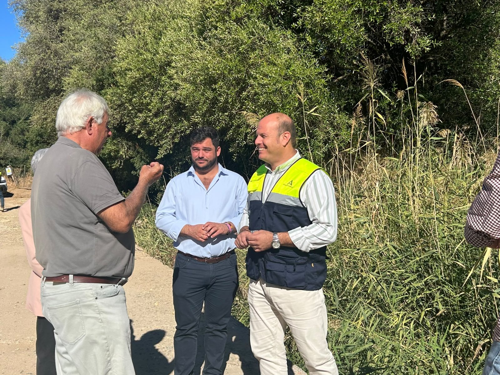 Tres hombres conversan de pie en un camino de tierra junto a vegetación alta y densa.