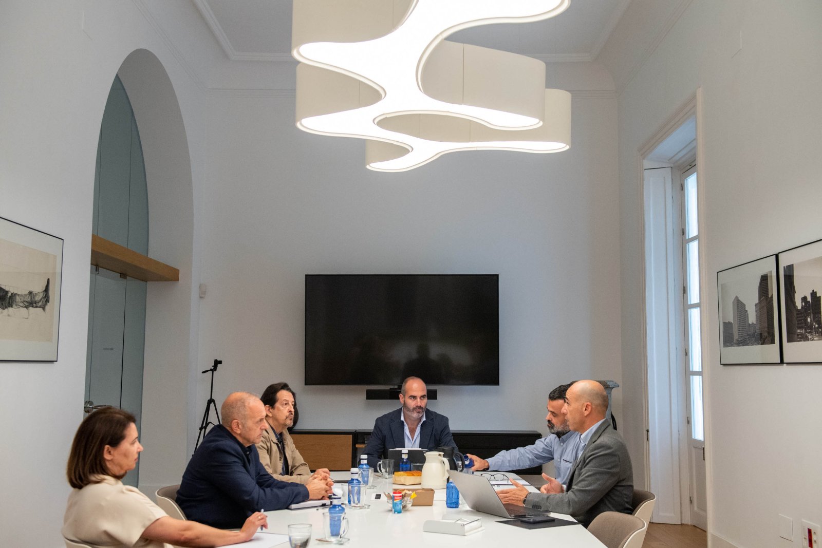 Seis personas sentadas alrededor de una mesa blanca de reuniones, con botellas de agua y un portátil, en una sala con una gran lámpara de techo y una pantalla a
