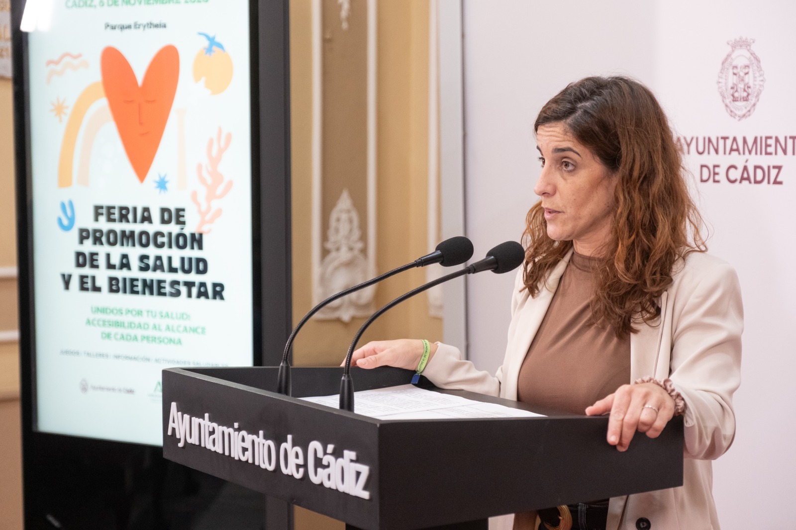 Una mujer habla ante dos micrófonos en un atril del Ayuntamiento de Cádiz, junto a una pantalla sobre una feria de salud.