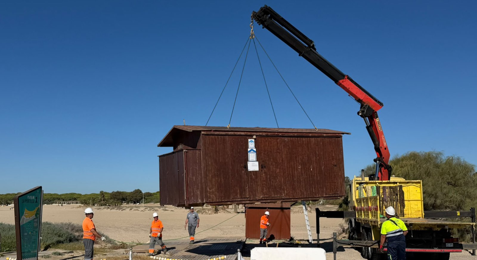 Una grúa eleva una caseta de madera sobre una playa mientras varios operarios con casco y chaleco observan la maniobra.