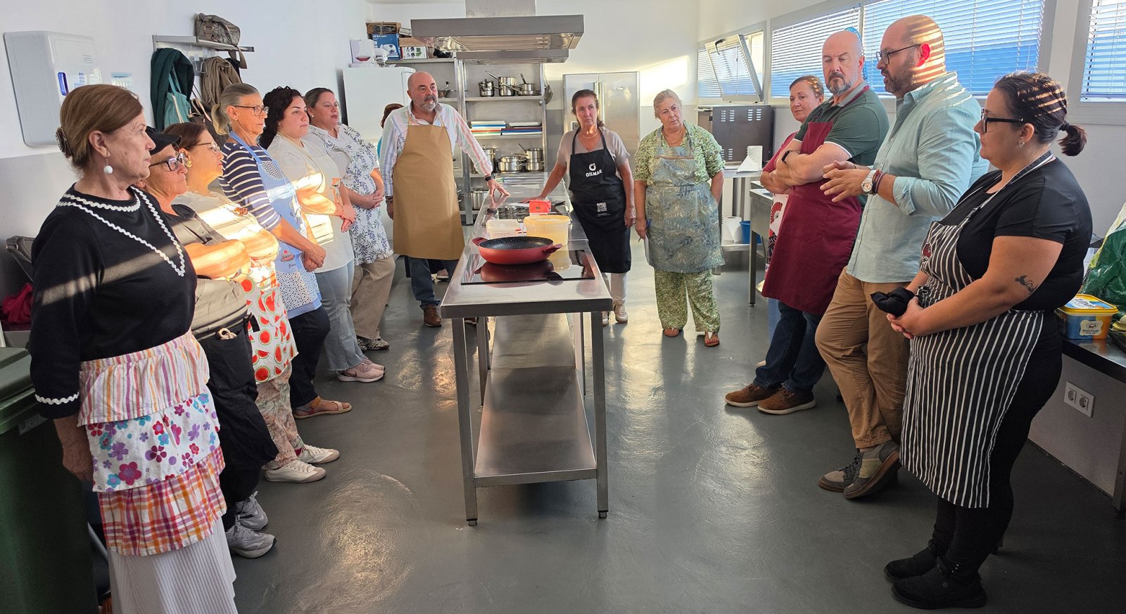 Un grupo de personas con delantales rodea una mesa de acero inoxidable en una cocina industrial.