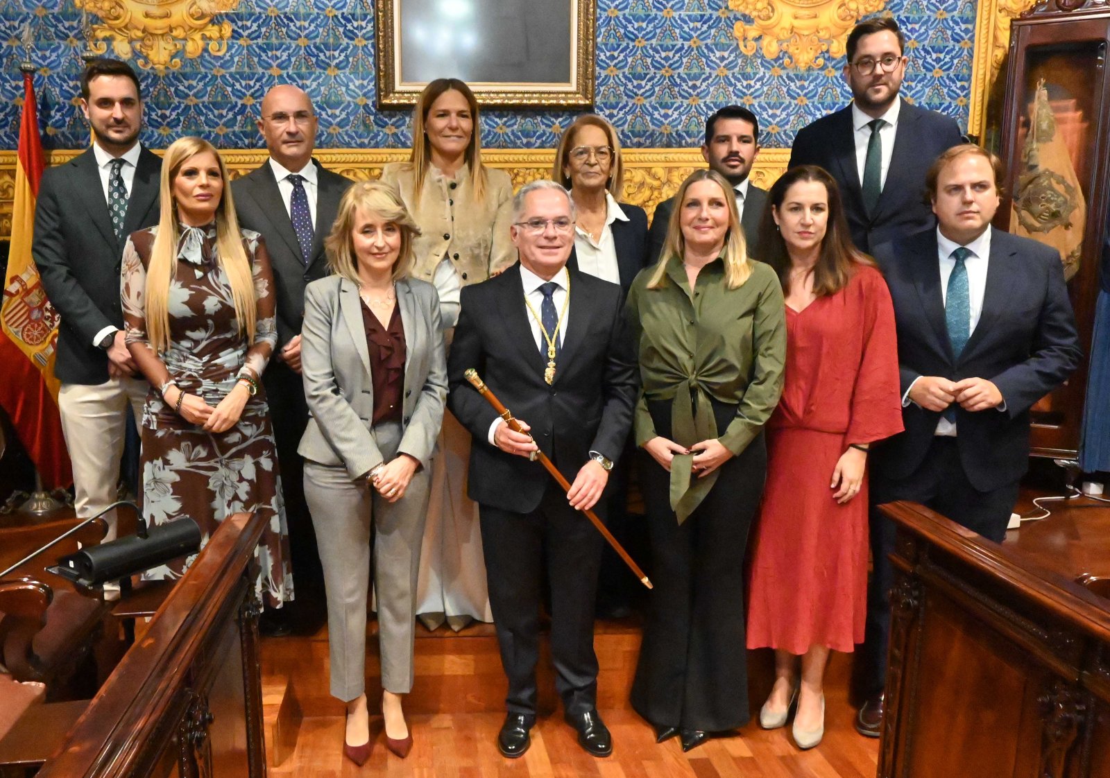Un grupo de once personas trajeadas posa de pie en una sala con paredes decoradas y un hombre sostiene un bastón de mando.