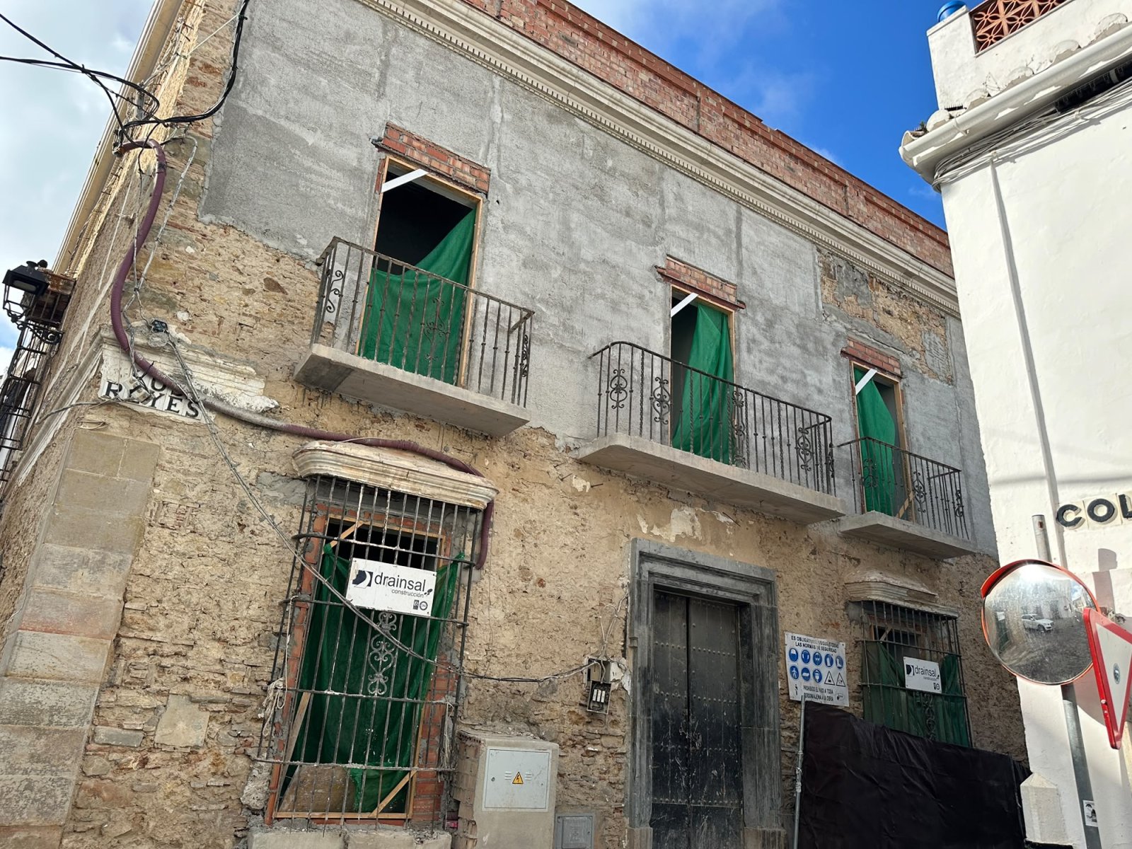 Fachada de un edificio antiguo en obras, con pared desconchada, tres balcones con lonas verdes y una puerta oscura.