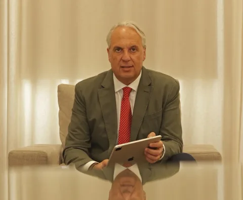 Un hombre mayor de traje verde y corbata roja, sentado con una tableta en la mano ante una mesa de cristal.