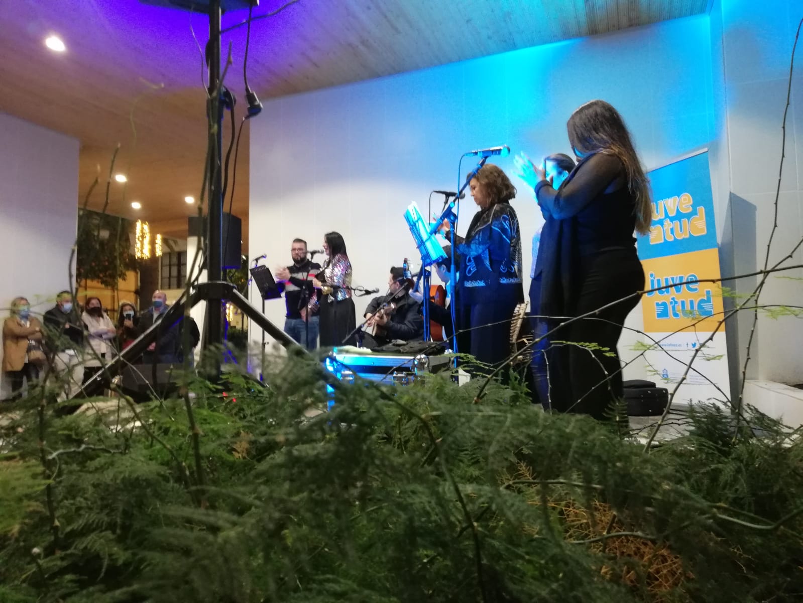 Un grupo de personas canta y toca instrumentos ante micrófonos en un escenario con luz azul, visto tras ramas verdes.