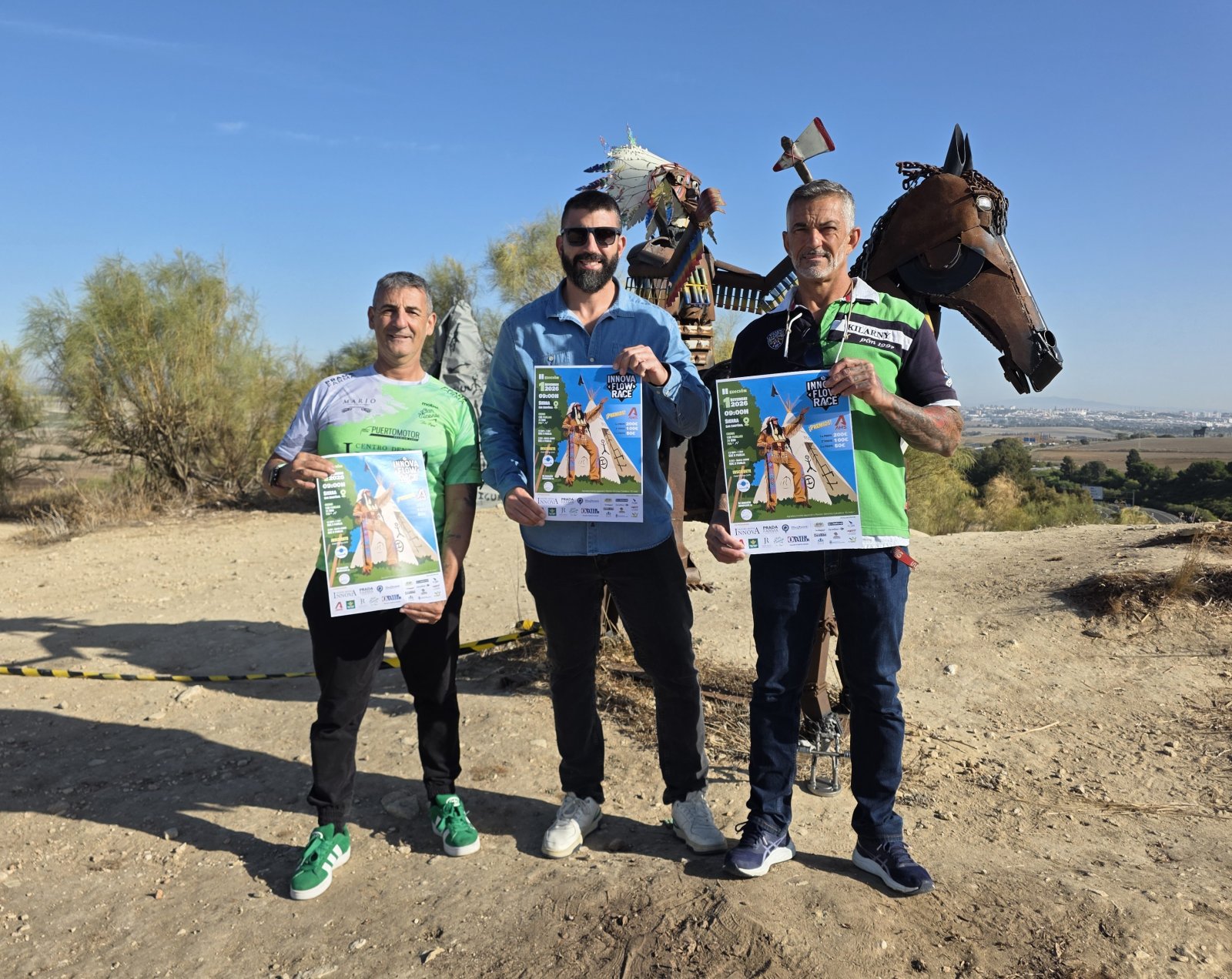 Tres hombres de pie al aire libre sostienen carteles con la inscripción Innova Flow Race ante una estructura metálica con forma de caballo.