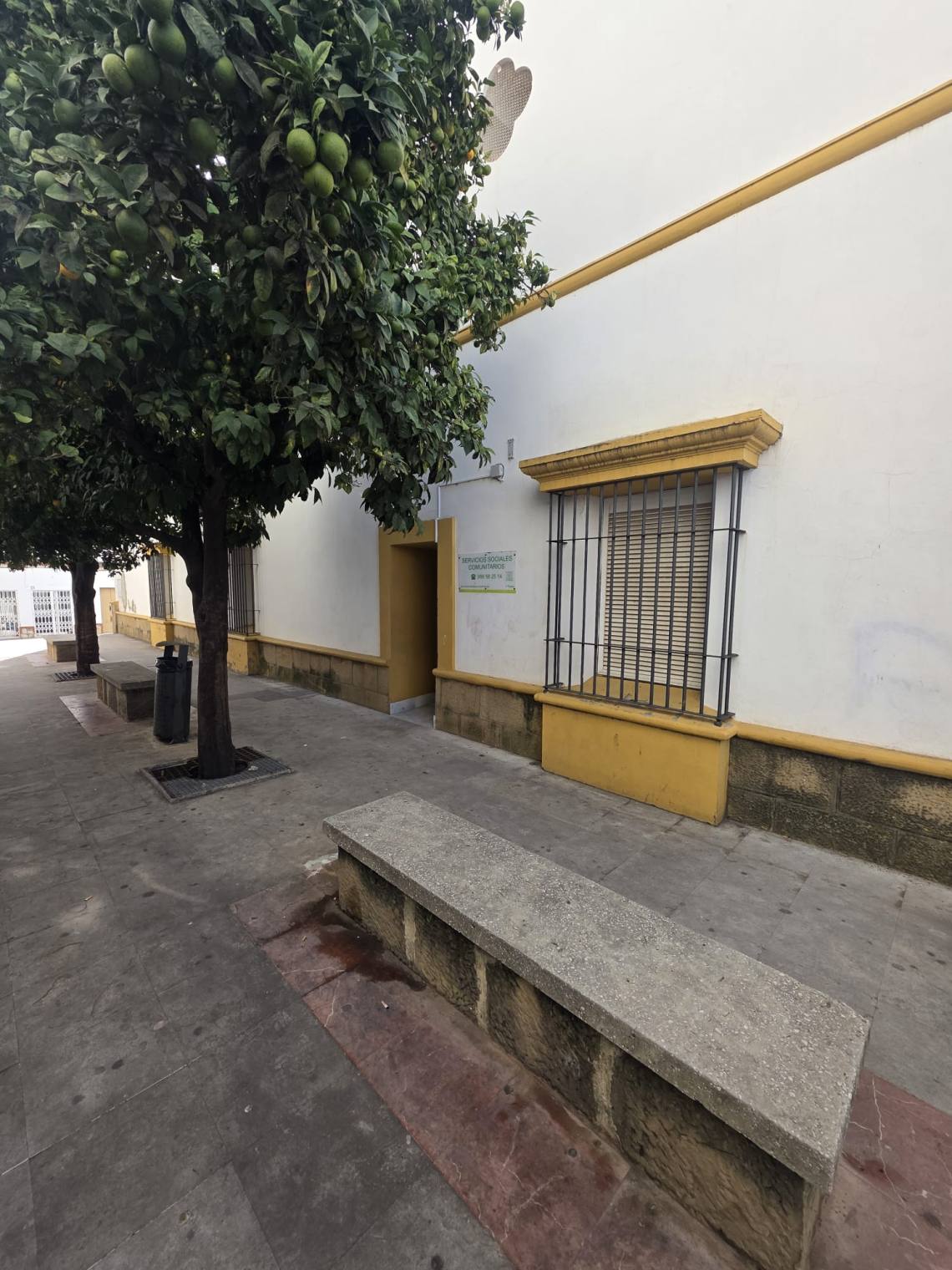 Fachada blanca con zócalo amarillo, ventana con reja y un cartel de servicios sociales, junto a naranjos y bancos en una acera.