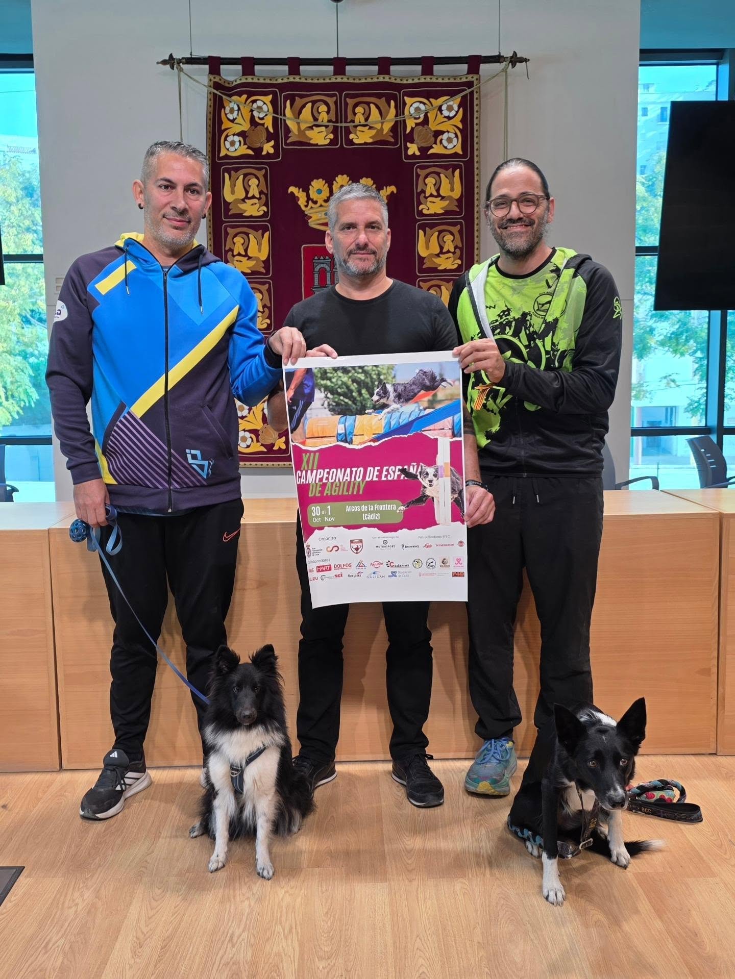 Tres hombres de pie sostienen un cartel de un campeonato de agility, con dos perros sentados delante.