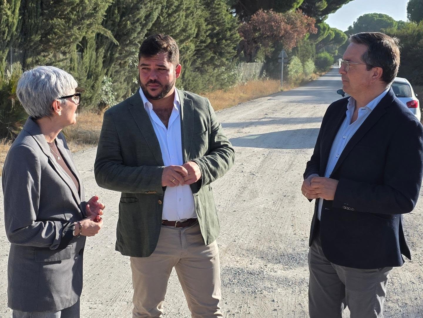 Tres personas con ropa formal conversan de pie en un camino rural de tierra, con árboles y un coche al fondo.