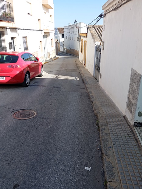 Una calle estrecha y cuesta arriba, con un coche rojo aparcado a la izquierda, fachadas blancas y una acera enlosada a la derecha.