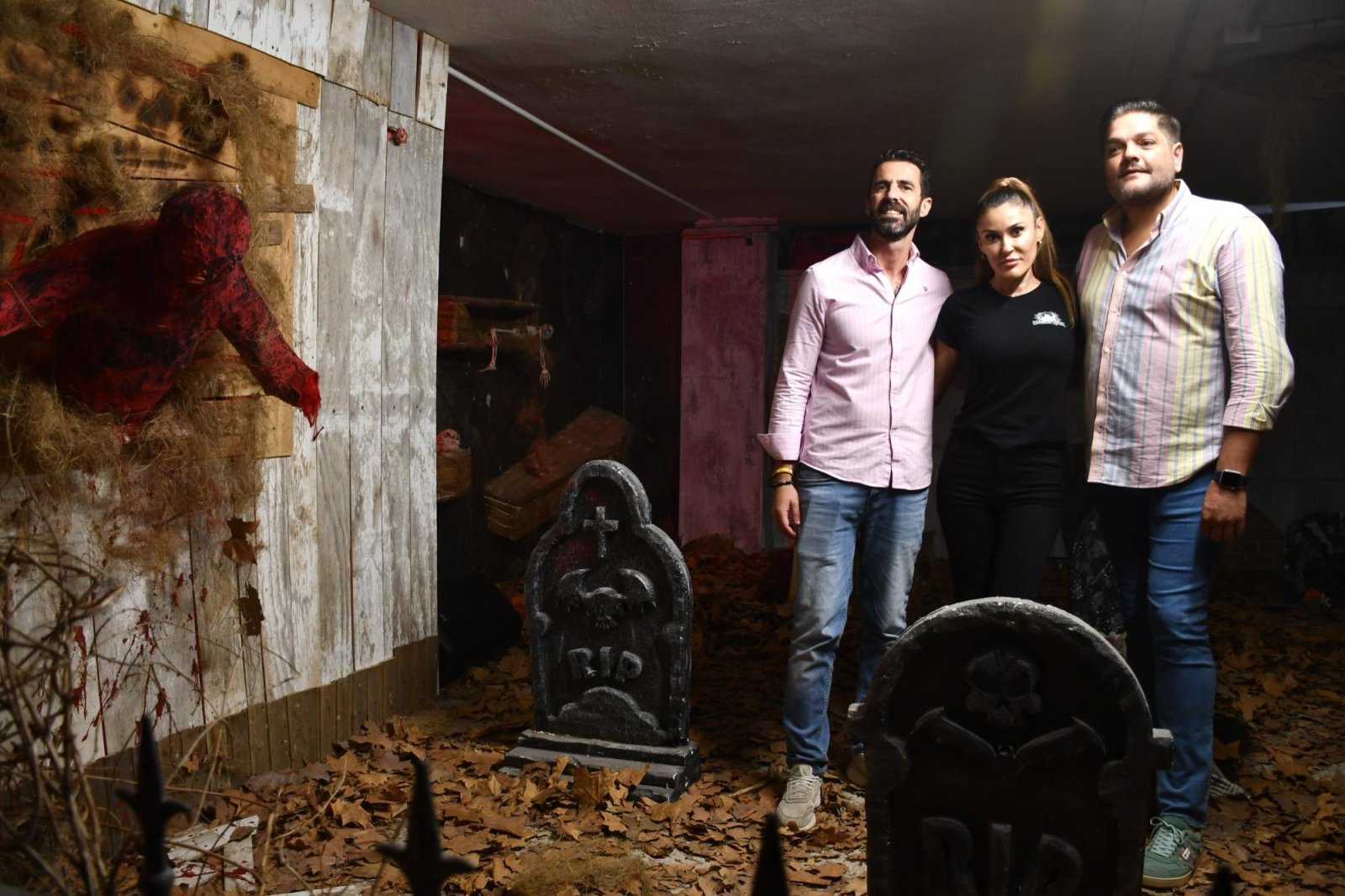 Tres personas de pie en un pasillo decorado de terror, con lápidas, hojas secas y un muñeco rojo colgado en una pared de madera.