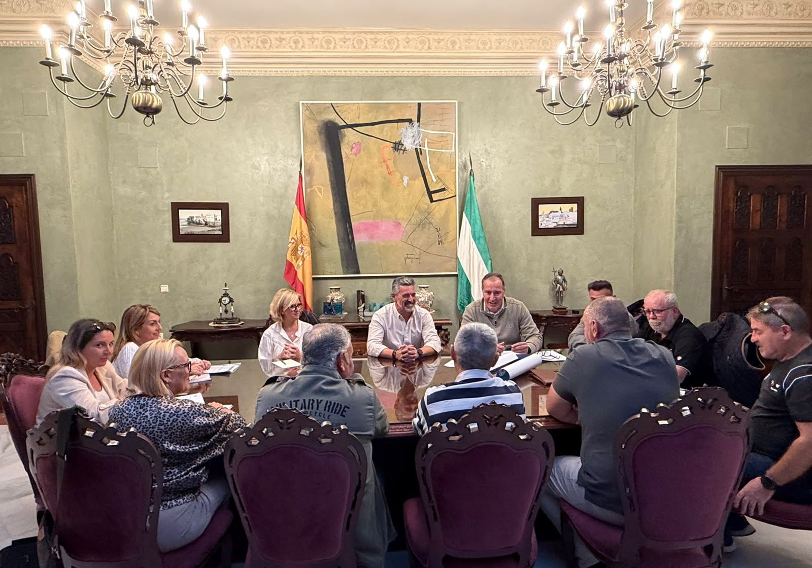 Un grupo de personas sentadas alrededor de una mesa de reuniones en una sala con lámparas de araña, banderas y un cuadro grande.
