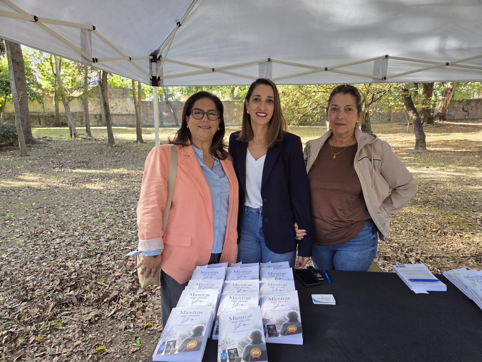 Tres mujeres de pie tras una mesa con ejemplares de un libro bajo una carpa blanca, en un parque.