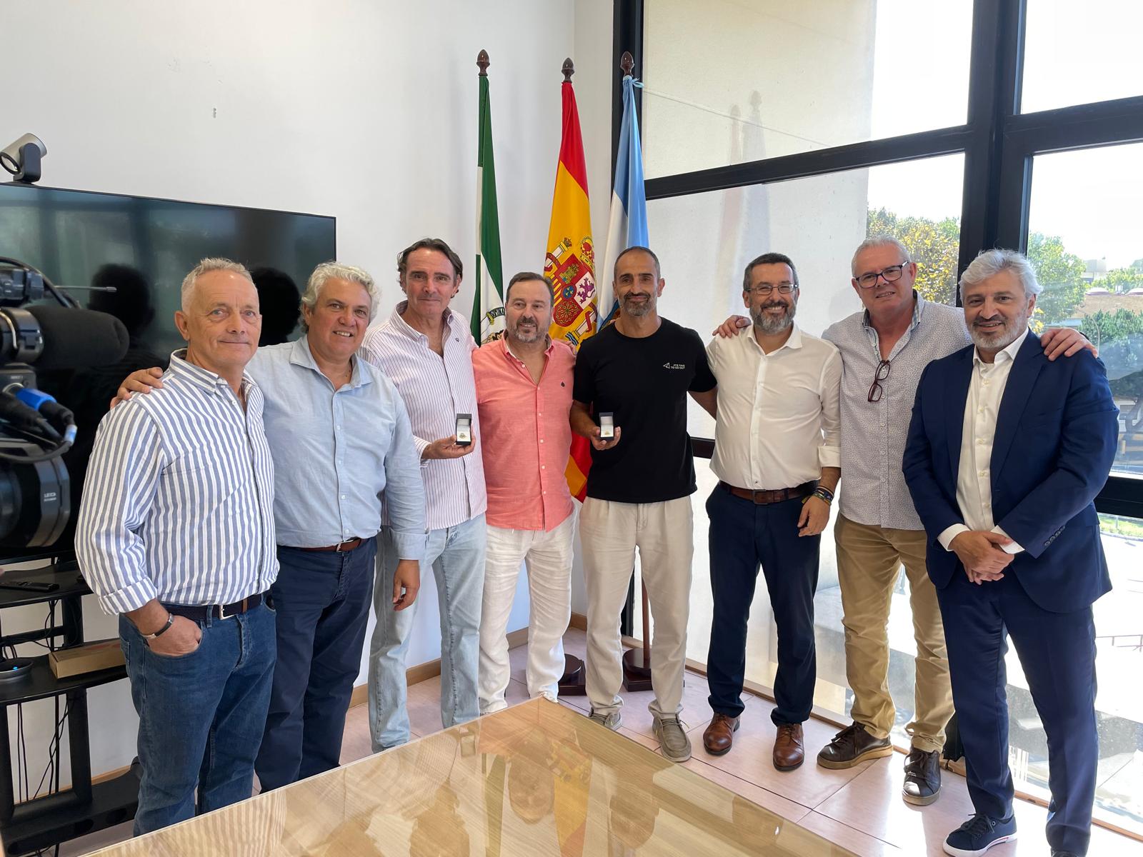 Ocho hombres posan de pie junto a una mesa, ante banderas y una ventana, en una sala.