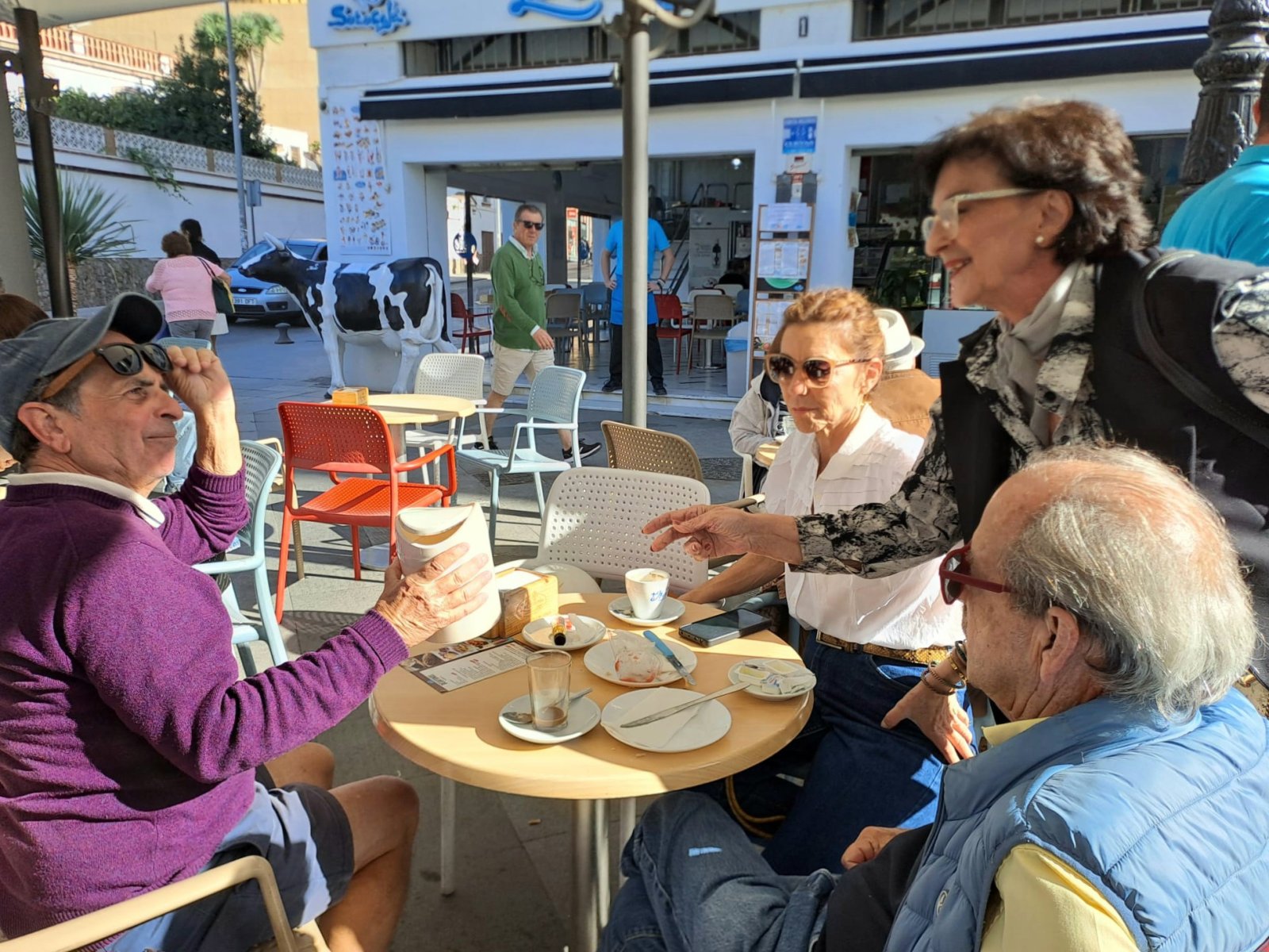 Cuatro personas mayores conversan sentadas en una mesa de terraza al aire libre, con tazas y platos, junto a una escultura de vaca.