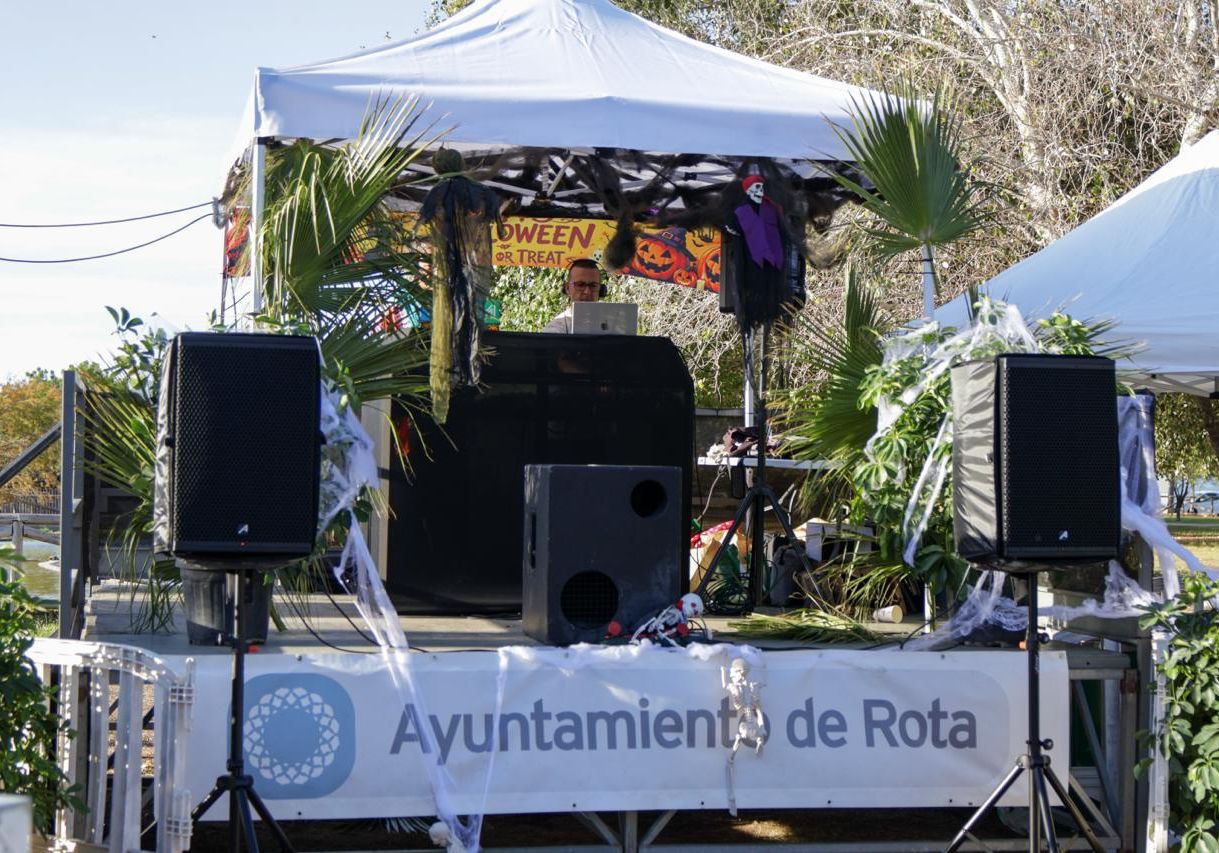 Escenario al aire libre con dos altavoces, carpa blanca, decoración de Halloween y una persona tras una mesa de mezclas.