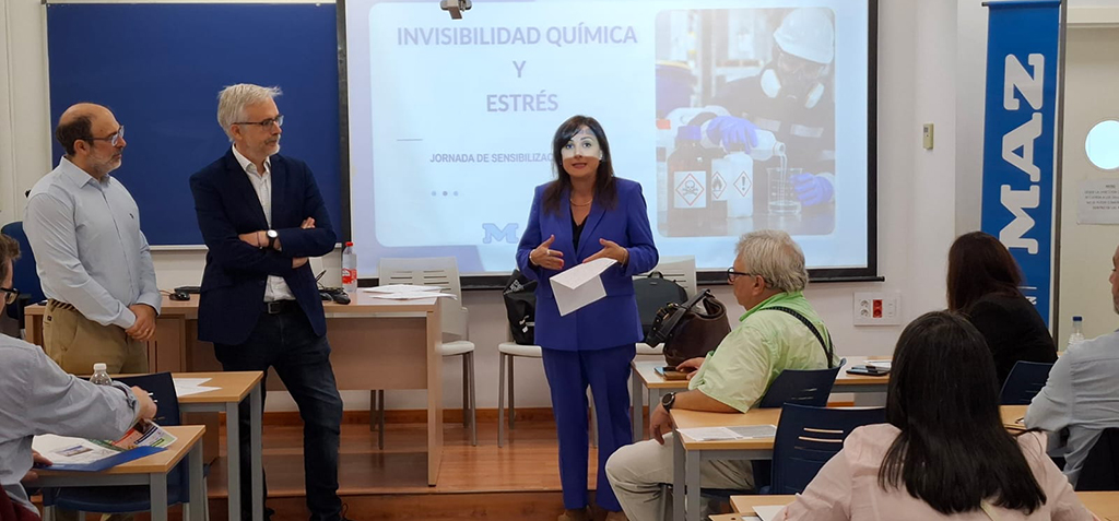 Tres ponentes junto a una pantalla con la proyección Invisibilidad química y estrés ante un público sentado en mesas de aula.