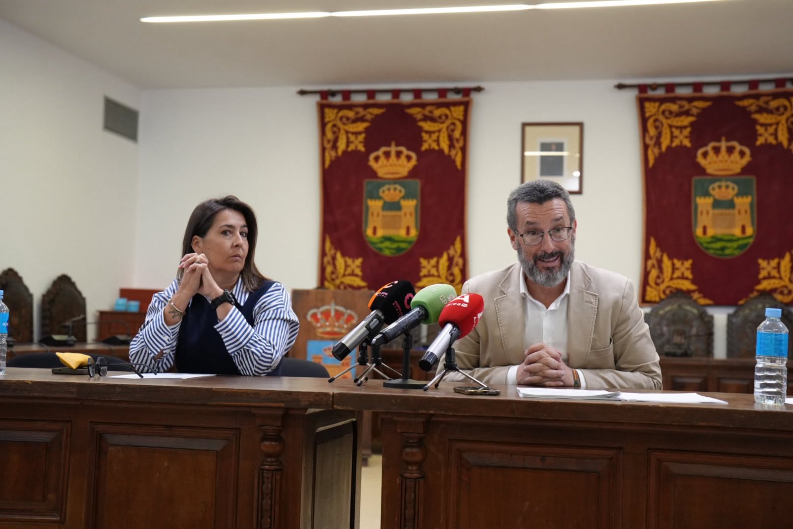 Un hombre con gafas y barba y una mujer con camisa de rayas están sentados tras una mesa con micrófonos en una sala con dos tapices.