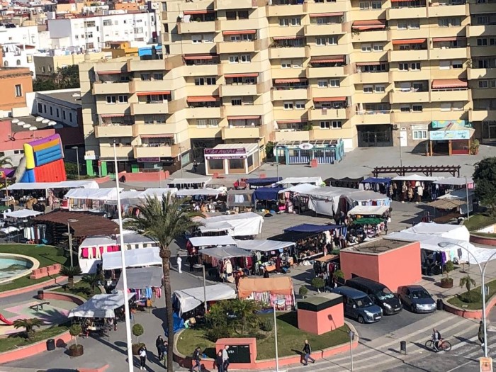 Vista elevada de un mercadillo al aire libre con muchos puestos de toldos blancos y azules ante grandes bloques de pisos amarillos.