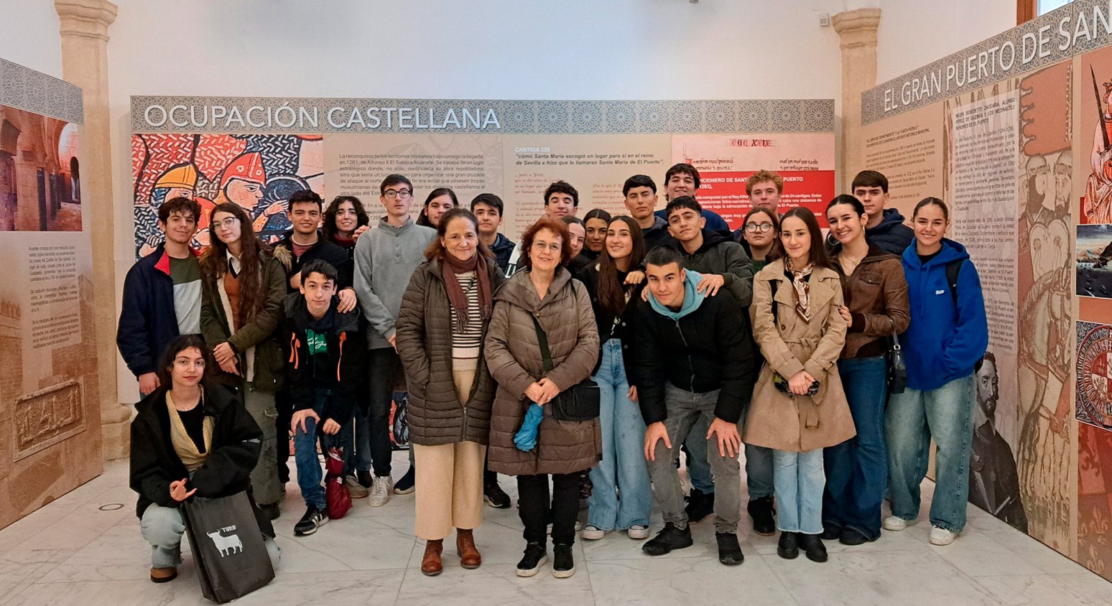Un grupo de jóvenes y dos mujeres adultas posan juntos ante paneles expositivos sobre la ocupación castellana en una sala de museo.