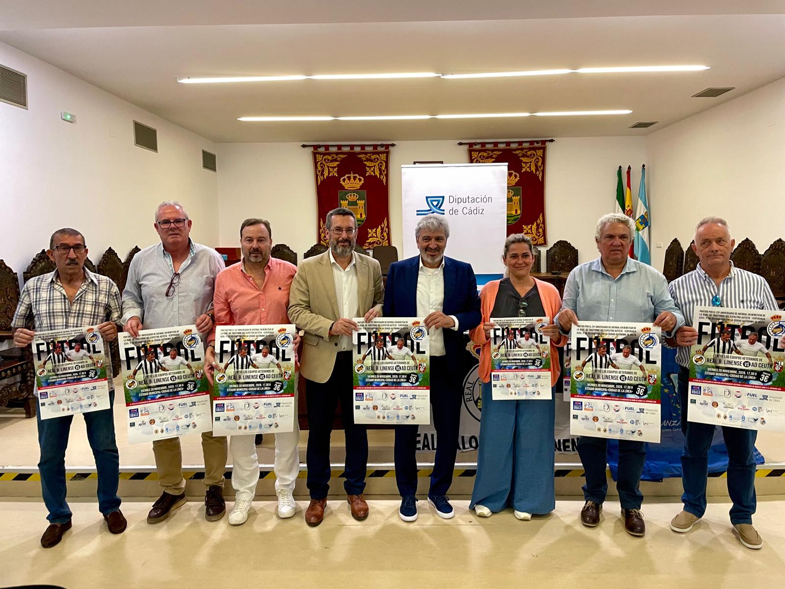 Siete personas de pie sostienen carteles de un partido de fútbol en una sala con banderas y un cartel de la Diputación de Cádiz.
