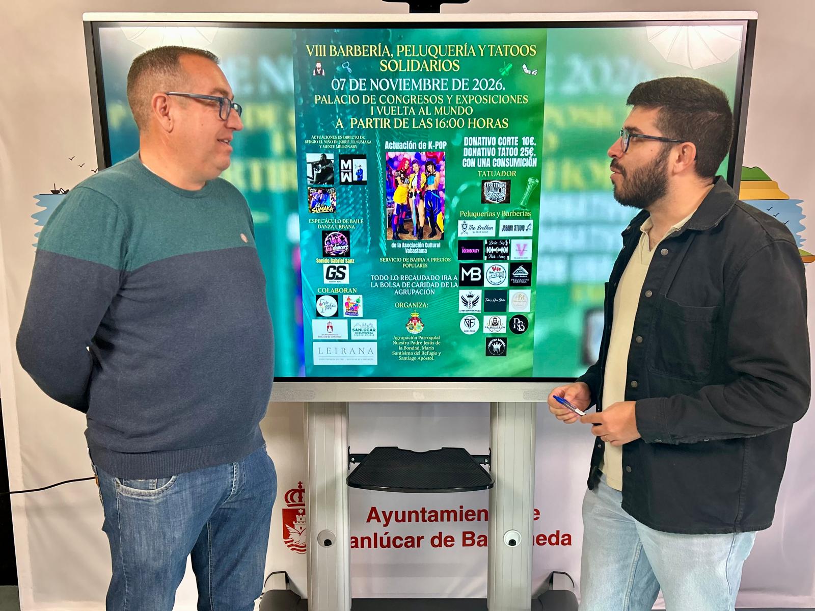 Dos hombres de pie conversan ante una pantalla grande que muestra un cartel verde de una jornada solidaria de barbería y tatuajes.
