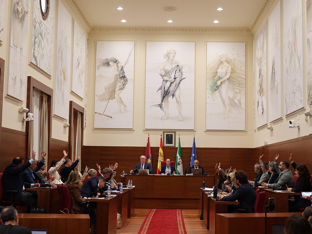 Sala de plenos con varias personas sentadas en escaños levantando la mano y tres figuras al frente ante banderas y grandes pinturas murales.